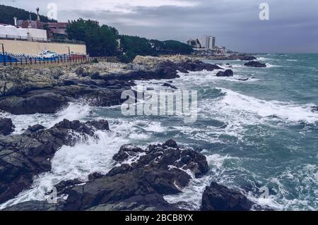Nahaufnahme von stürmischen Wellen und übergiebeltem Himmel mit dem Stadtbild der Küste von Busan Stockfoto