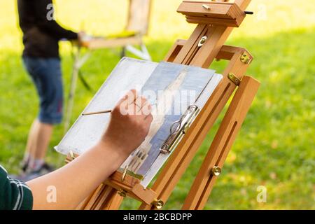 Zwei Mädchen Künstler malen auf freiem Himmel, Holz-Staffelei Skizzenbuch mit unvollendeter Malerei Nahaufnahme. Weibliche Hand malt auf einer sonnigen Sommerwiese mit Öl Stockfoto