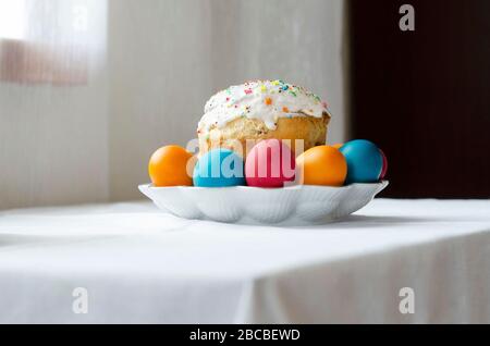 Weiße Platte mit einem glasierten osterkuchen und bemalten Eiern stehen auf einem Tisch mit einem weißen Tuch bedeckt. Vorbereitung auf die Osterfeier. Stockfoto