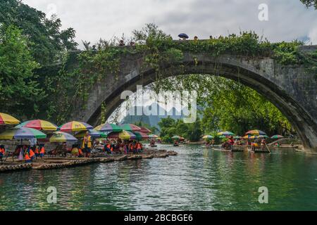 Yangshuo, China - August 2019: Kleine lokale touristische Passagier Bambus boote Menschen, die im Rahmen der historischen Yulong Qiao Brücke auf der Reise entlang. Stockfoto