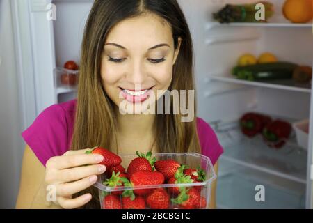 Nahaufnahme des hübschen lächelnden Mädchens in der Nähe des Kühlschranks und Blick auf Erdbeeren. Gesundes Lebensmittelkonzept. Frisches Obst und Gemüse. Stockfoto