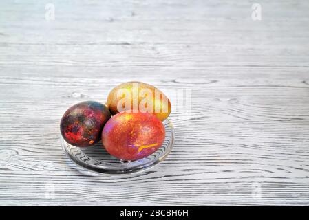 Drei farbige Eier in einer transparenten Untertasse auf einem hellen natürlichen Hintergrund mit einer uneinheitlichen Struktur Stockfoto