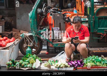Zigong, China - Juli 2019: Chinesin sitzt auf dem Straßenpflaster und verkauft Gemüse, Zigong Stadt, Provinz Sichuan Stockfoto