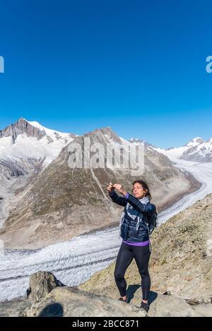 Junge Kaukasierin mit Rucksack auf dem Rücken, die ein Selfie mit dem monumentalen Aletschgletscher hinter sich macht. Stockfoto