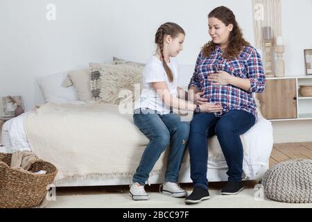 Tochter vor Teenager, die den Bauch ihrer schwangeren Mutter berührt, Leute, die auf einem breiten Bett im Schlafzimmer sitzen Stockfoto