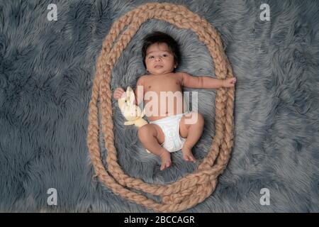 Porträt eines neugeborenen Jungen, Alter 1 Monat, in einer Windel, schwarzes Haar, blaue Augen auf grauen Pelzdecke mit einem Teddybären und lächelnd. Familie, Liebe, ch Stockfoto
