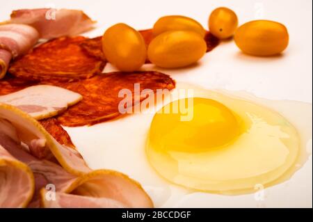 Ein zerbrochenes Hühnerei ohne Schale, Speck- und Wurstscheiben sowie Tomaten auf weißem Grund. Nahaufnahme Stockfoto