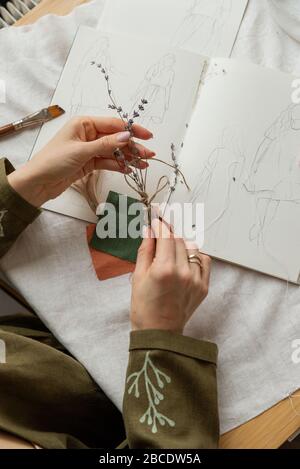 Über dem Blick auf weibliche Künstler, die trockene Lavendelblüten mit Händen in der Nähe kreativer Skizzen von Leinenkleid halten. Flacher Arbeitsplatz von Seestrees, Arbeit Stockfoto