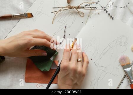 Blick von oben auf die Hände des Designers, der Skizzen zukünftiger Leinenkleider macht und die Pinsel für den Zeichendruck hält. Arbeitsplatz von Seestrees mit Zitzen von o Stockfoto