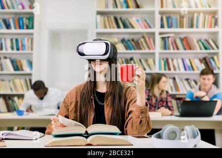 Nahaufnahme einer hübschen kaukasischen Frau in der Bibliothek, mit VR-Headset und neuer Technologie zum Studieren, Trinken von Kaffee und Spiegeln Stockfoto