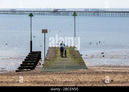 Southend-on-Sea, Großbritannien. April 2020. Während der COVID-19-Sperrung spaziert eine Einzelfigur mit dem Fahrrad entlang eines moosigen Stegs an der Küste von Southend. Grüne Navigationsmarkierungen und ferne Industriestrukturen bilden den Rahmen für die beschauliche Mündungsszene. Aufgenommen an einem warmen Frühlingstag unter sozialen Distanzierungsbeschränkungen. Penelope Barritt/Alamy Live News Stockfoto