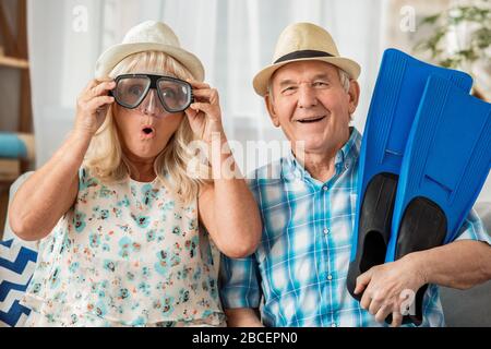 Ein älteres Paar sitzt im Reisebüro und trägt Hüte und eine Schwimmbrille, hält Flossen und schaut in die Kamera Stockfoto