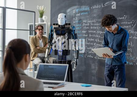 Cleverer Teenager-Typ mit Laptop, der an der Tafel vor der Klasse steht und Roboterfähigkeiten und -Eigenschaften präsentiert Stockfoto