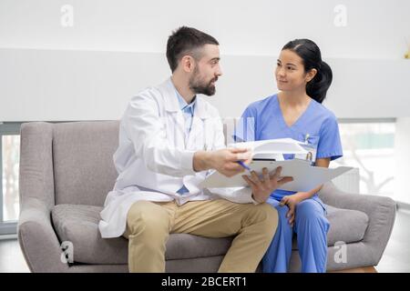 Der junge Arzt in Whitecoat erklärt seinem hübschen Assistenten in blauer Uniform die Informationen in einem Dokument, während er in der Lobby der Kliniken sitzt Stockfoto