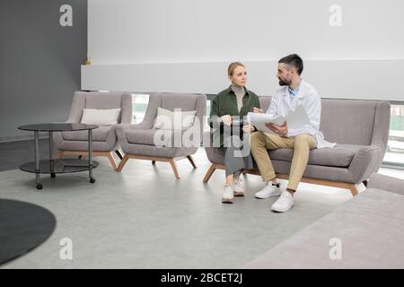 Ziemlich junge Patientin von Zahnkliniken, die ihren Zahnarzt aufsucht, der Informationen in einem medizinischen Dokument erklärt, während beide in der Lounge sitzen Stockfoto