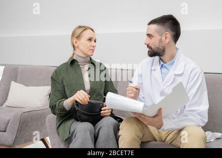 Junger, selbstbewusster Arzt, der Notizen macht, während er einen seiner Patienten in der Lounge von Zahnkliniken berät Stockfoto
