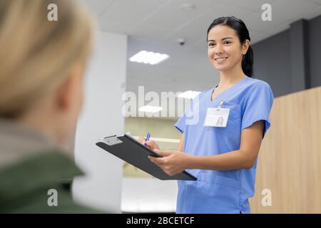 Junger lächelnder Assistent in blauer Uniform macht sich Notizen in der Registrierungsunterlage, während er sich einen der Patienten von Zahnkliniken ansieht Stockfoto