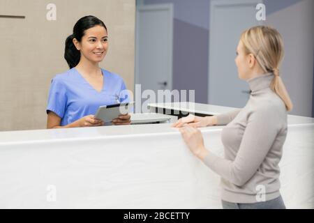 Ziemlich junge Beraterin mit Tablette am Rezeptionsschalter vor der weiblichen Patientin und Registrierung für einen medizinischen Termin Stockfoto