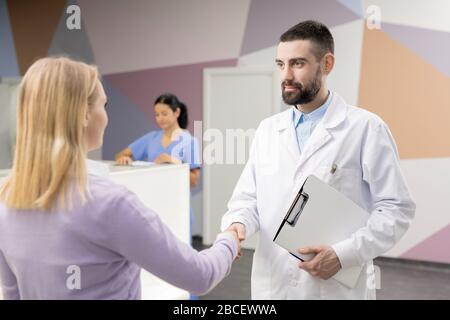 Junger selbstbewusster Arzt im Whitecoat-Handshake mit einem der Patienten im Hintergrund des Rezeptionszählers und seinem Assistenten in Uniform Stockfoto