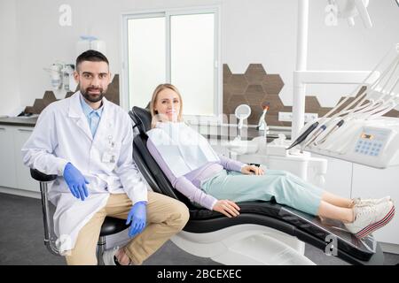 Glückliche blonde Patientin und junge erfolgreiche Zahnärztin im Whitecoat und Handschuhe sitzen im Zahnarztbüro zeitgenössischer Kliniken Stockfoto