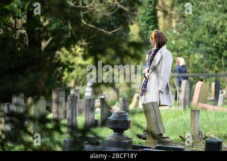 Eine junge Frau, die einen grauen Mantel trägt und an einem sonnigen Tag unter Grabsteinen auf einem Friedhof nachdenklich steht. GROSSBRITANNIEN Stockfoto