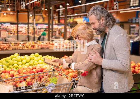 Reifes liebevolles Paar in der Casualwear, das auf Notizblock Seite mit Einkaufsliste schaut, während er sich auf dem modernen Hypermarkt bewegt Stockfoto