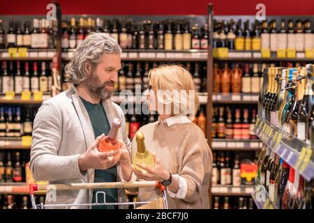 Glückliches Paar im Alter mit Einkaufswagen, das Wein auswählt, während es in einer der Supermarktabteilungen unter den Regalen mit Alkohol steht Stockfoto