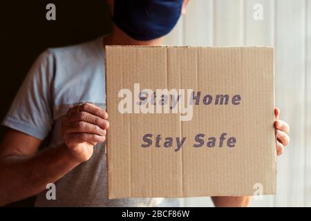 Eine Person, die Schutzmaske trägt, die Hand hält ein Schild, das zeigt, dass sie zu Hause bleibt, für Selbstisolation und soziale Entfernungen sicher ist. Hintergrundkonzept für Corona Stockfoto