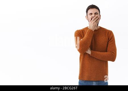 Portrait des müden bärtigen Mannes fühlt sich nach der Arbeit in der späten Nacht erschöpft, gähnelt und bedeckt Mund mit Handfläche, steht gelangweilt oder schläfrig, wartet auf Tasse Stockfoto