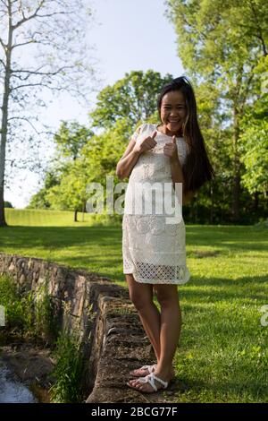 Ein schönes junges Filipina-Mädchen in Weiß gibt das Daumen-up-Zeichen, während sie für ihre Senior Pictures in Sunken Gardens in Huntington, Indiana, USA posiert. Stockfoto