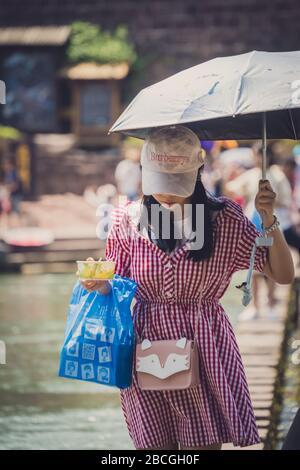 Feng Huang, China - August 2019: Mädchen der Chinesin, die Regenschirm hält und Essen und Bagwalking auf den Trittsteinen über den Fluss Tuo, flo, wegnehmen Stockfoto