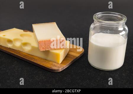 Emmentaler Käse und Gruyere Käse auf einem Holzbrettstand neben einem Glas mit Milch auf einem schwarzen Tisch. Stockfoto