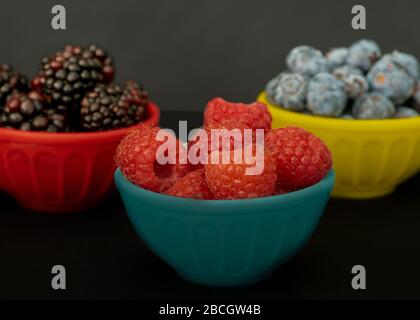 Brombeeren, Blaubeeren und Himbeeren in bunten Schalen von der Seite betrachtet, auf schwarzer Oberfläche und Hintergrund, gesundes Esskonzept Stockfoto