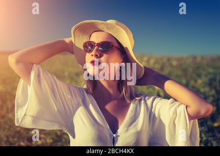 Junge Frau porträtiert im Feld in weißem Kleid und trägt einen Hut und eine Sonnenbrille Stockfoto