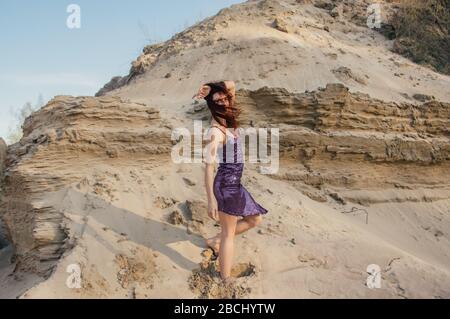 Braunhaarige Frau in violett glitzerndem Kleid, die auf Sandhang geht Stockfoto