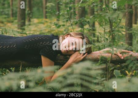 Porträt des jungen schönen Mädchens in schwarzem Kleid im Wald, das auf dem Baumstamm liegt, geschlossene Augen Stockfoto