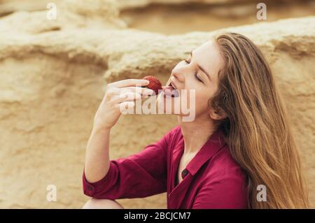 Wunderschönes blondes lächelndes Mädchen im violetten Kleid, das frische Erdbeere auf die Natur leckt Stockfoto