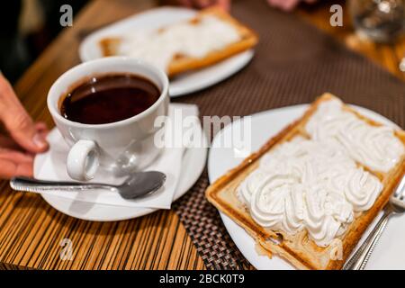 Nahaufnahme Makroansicht von Hand, die geschmolzene heiße Schokoladenlöffeltasse und belgische Waffel auf weißem Teller mit Gabel, die morgens mit Schlagsahne bekrönt wurde Stockfoto