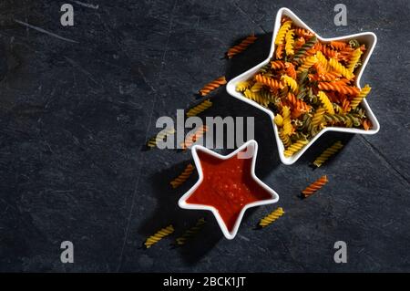 Konzeptzusammensetzung von bunten Fusilli-Pasta und Tomatensauce in sternförmiger Schüssel auf dunklem Schiefergrund. Stockfoto