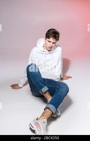 Modisches, gutaussehendes, junges europäisches Männermodell, bekleidet mit weißem Sweatshirt, blauer Jeans und weißen Turnschuhen, die im Studio auf einem pinkfarbenen Hintergrund für posieren Stockfoto