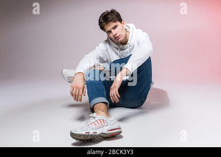 Modisches, gutaussehendes, junges europäisches Männermodell, bekleidet mit weißem Sweatshirt, blauer Jeans und weißen Turnschuhen, die im Studio auf einem pinkfarbenen Hintergrund für posieren Stockfoto