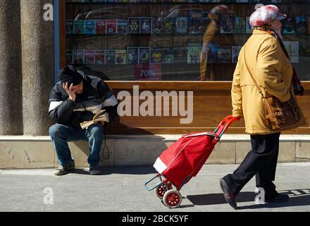 Bukarest, Rumänien - 16. März 2020: Eine ältere Frau mit einem Einkaufswagen geht an einem Mann vorbei, der in der Nähe einer Buchhandlung sitzt und ein Buch liest. Stockfoto