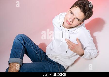 Modisches, gutaussehendes, junges europäisches Männermodell, bekleidet mit weißem Sweatshirt, blauer Jeans und weißen Turnschuhen, die im Studio auf einem pinkfarbenen Hintergrund für posieren Stockfoto