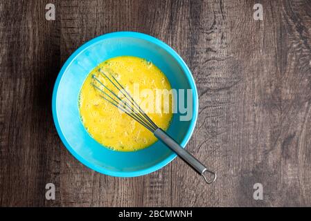 Gepeitschte Eier in blauer Mischschale, Metall-Schneebesen, Holztisch Stockfoto