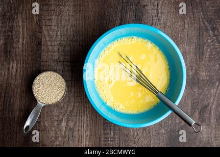 Gepeitschte Eier in blauer Mischschale, Metall-Schneebesen, Messbecher mit rohem Quinoa, Holztisch Stockfoto