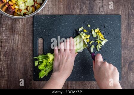 Die Hände der Frau hacken die Stangen des Sellerie mit dem Küchenmesser auf schwarzem Schneidebrett Stockfoto