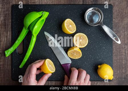 Die Hände der Frau schneiden eine Zitrone auf einem schwarzen Schneidebrett, Küchenmesser, grüne Zitruspressemaschine, Messbecher Stockfoto