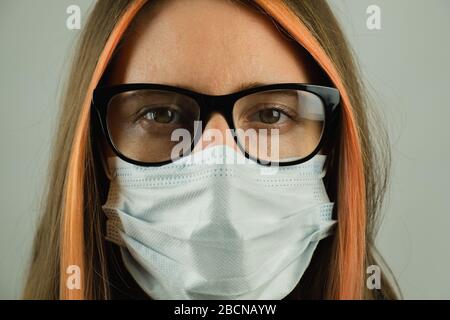 Intelligente Frau in Brillen mit medizinischer Maske, Nahporträt mit Fokus auf Augen. Junge Frau in chirurgischer Maske in dunkelgrauer Kulisse. Konce Stockfoto