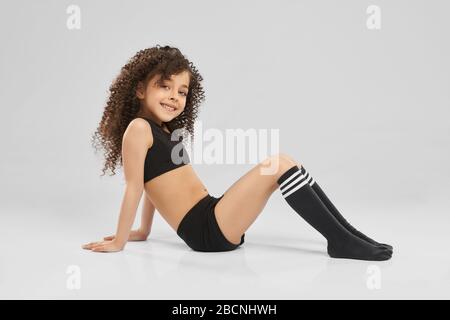 Seitenansicht von liebenswert lächelnd Mädchen in schwarzer Sportkleidung und Kniestrümpfe posiert, während sitzen vom Boden, isoliert auf grauem Hintergrund. Wenig professionell Stockfoto