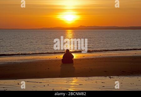 Portobello, Edinburgh, Schottland, Großbritannien. 5.. April 2020. Sonnenaufgang am Strand von Portobello, mit einer Frau, die am Sandstrand in Kontemplation sitzt, und verschiedenen anderen Menschen, die Schwimmen, Wandern, Laufen fotografiert haben. Stockfoto